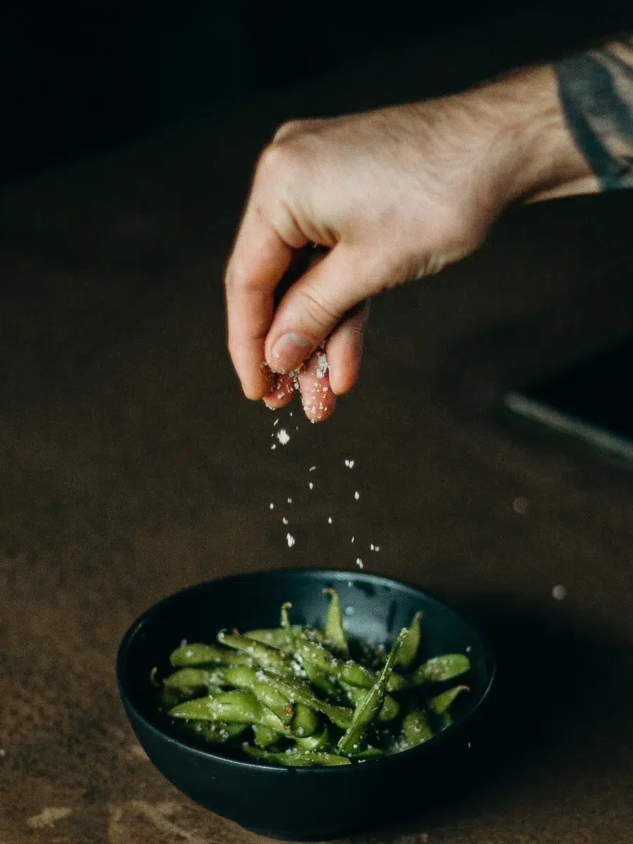 Eine Hand streut Salz Ã¼ber eine dunkle Schale mit Edamame